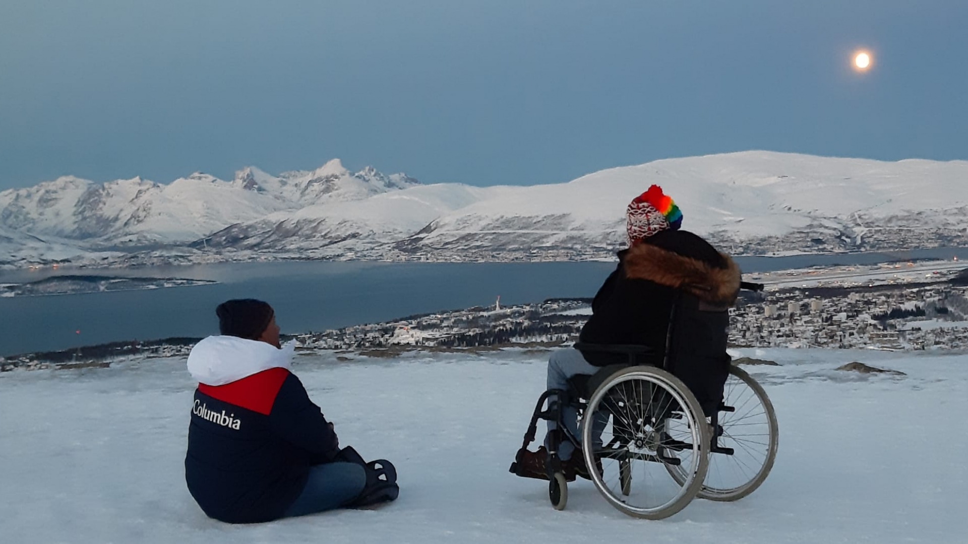 Person sitting in a wheelchair with another person close by, phototaken from the cable car with view over Tromsøya and Kvaløya