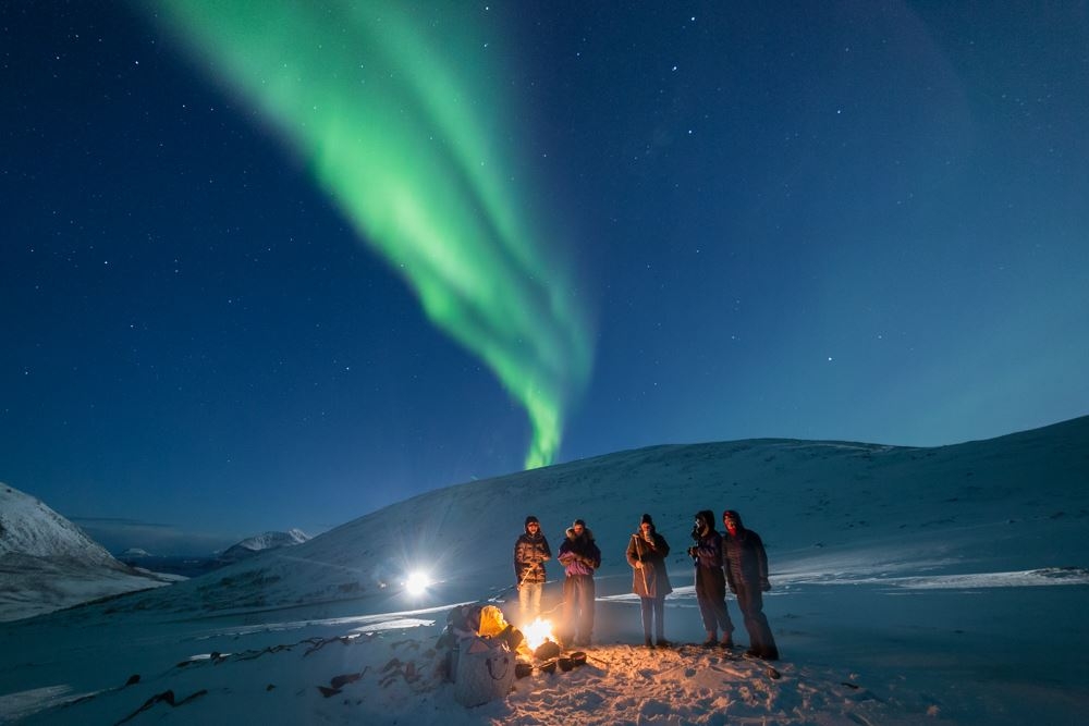 Persons standing under the norther lights around a bonfire 