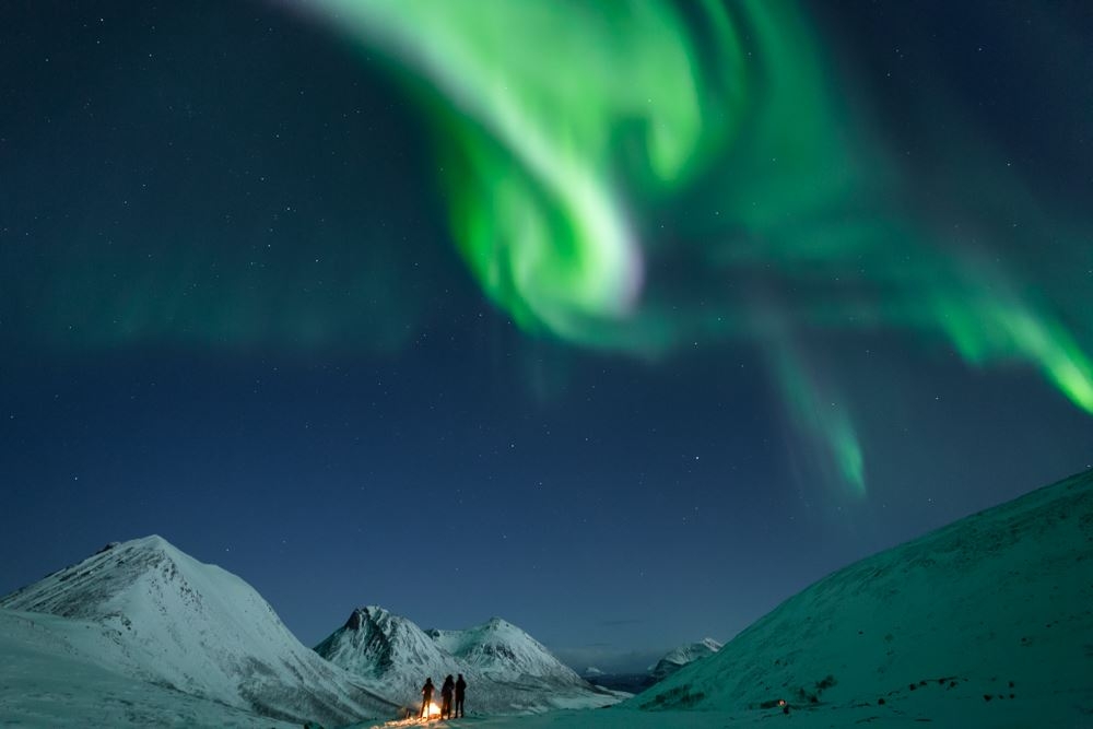 Multiple people standing under the norther lights around a bonfire with snow covered surrounding mountains 
