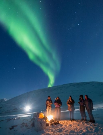 Persons standing under the norther lights around a bonfire 