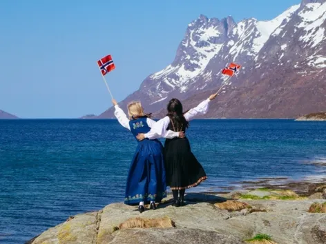 Two friends dressed in bunad standing by the ocean holding the Norwegian flag.