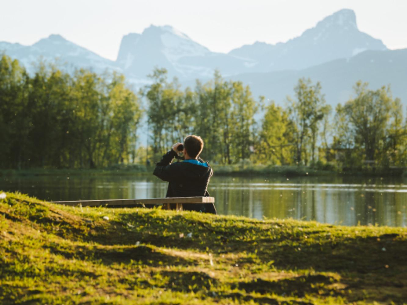 En person sitter på en benk og ser i en kikkert på fjellene i bakgrunnen.