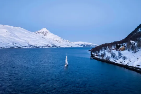 Sailboat on calm winter water surrounded by snow-covered mountains and trees, with a house by the shoreline