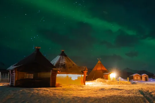 Northern lights over snow-covered cabins and a lavvu lit by warm lights.