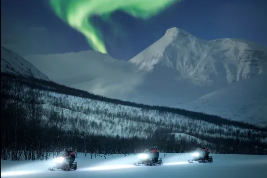 Guests drive snow scooters with the Aurora above their heads