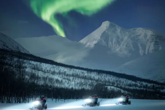 Guests drive snowmobiles past the mountains and the shining Northern Lights