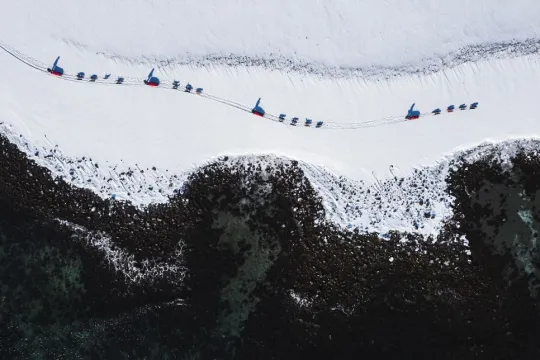 Dog sled teams travelling through a snow-covered Arctic landscape, seen from above with dark mountain formations in the foreground.
Dog sledding