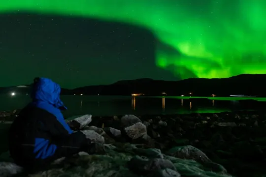 En gjest sitter ved fjorden og nyter synet av grønt nordlys over fjellene. En stemningsfull opplevelse under en guidet nordlysjakt i Tromsø-regionen.