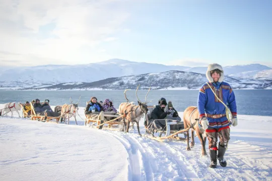 Reindeer pulling sleds with people across a snow-covered landscape in front of mountains and water.