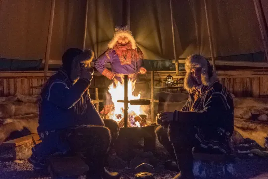 Three people in traditional clothing gathered around a campfire inside a tent.