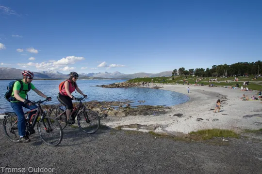 Two people cycling along a coastal path by the beach in Tromsø, with mountains and sea in the background.