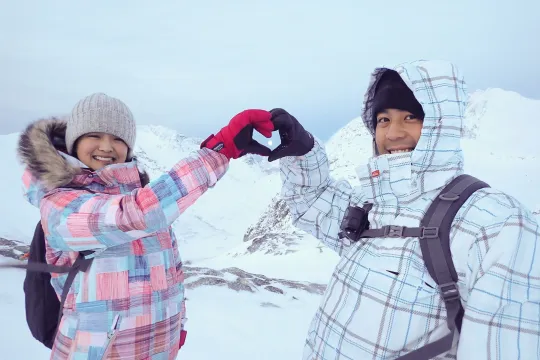 Two people in winter clothing forming a heart shape with their hands in snowy mountains.