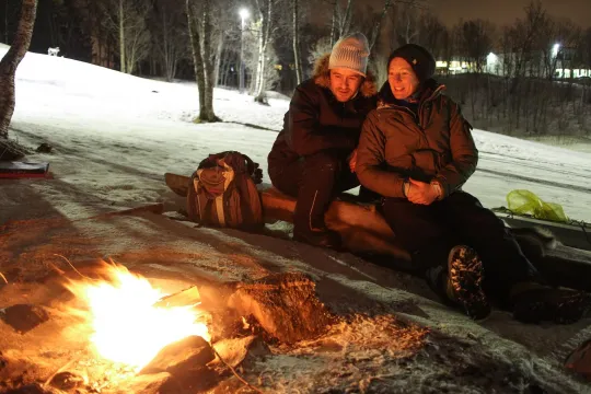 Two people by a campfire in a snowy landscape, with trees and a backpack in the background.