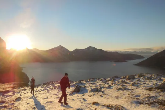 Two people walking in snowy mountain terrain with sunset and water in the background.