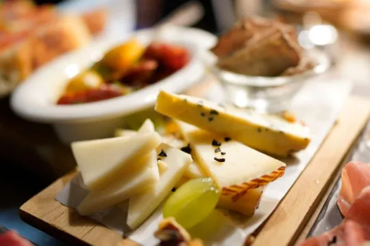 Nærbilde av ost med sorte frø, grønne druer og kjeks, med spekemat og bruschetta i bakgrunnen.