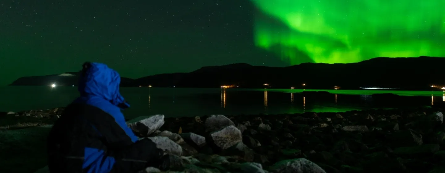 A guest in a blue winter jacket sits by a rocky shoreline, watching the green Northern Lights above a fjord near Tromsø.