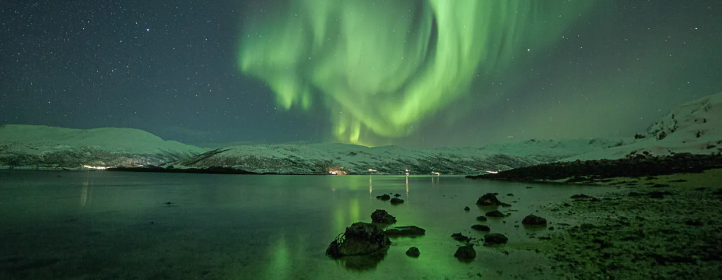 Grønt nordlys over strandlinjen, med snødekte fjell i bakgrunnen