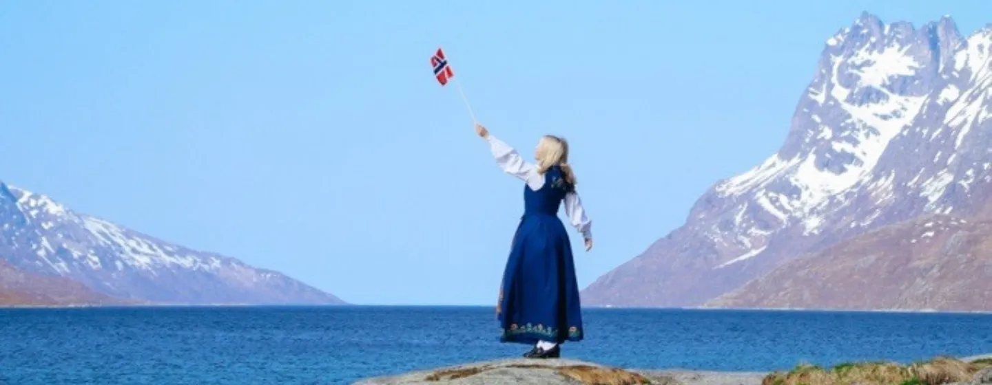 Dame i bunad som står ved sjøen og vifter med det norske flagget.